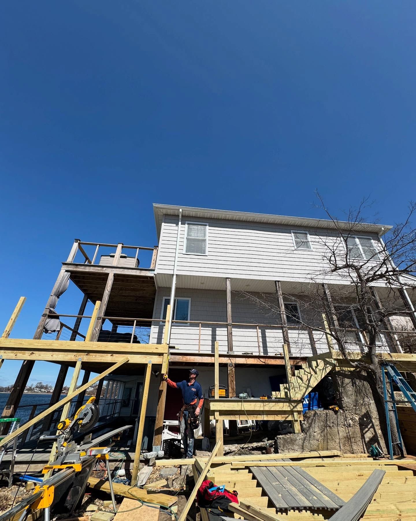 A person stands under a large, partially constructed wooden deck attached to the side of a two-story waterfront house.
