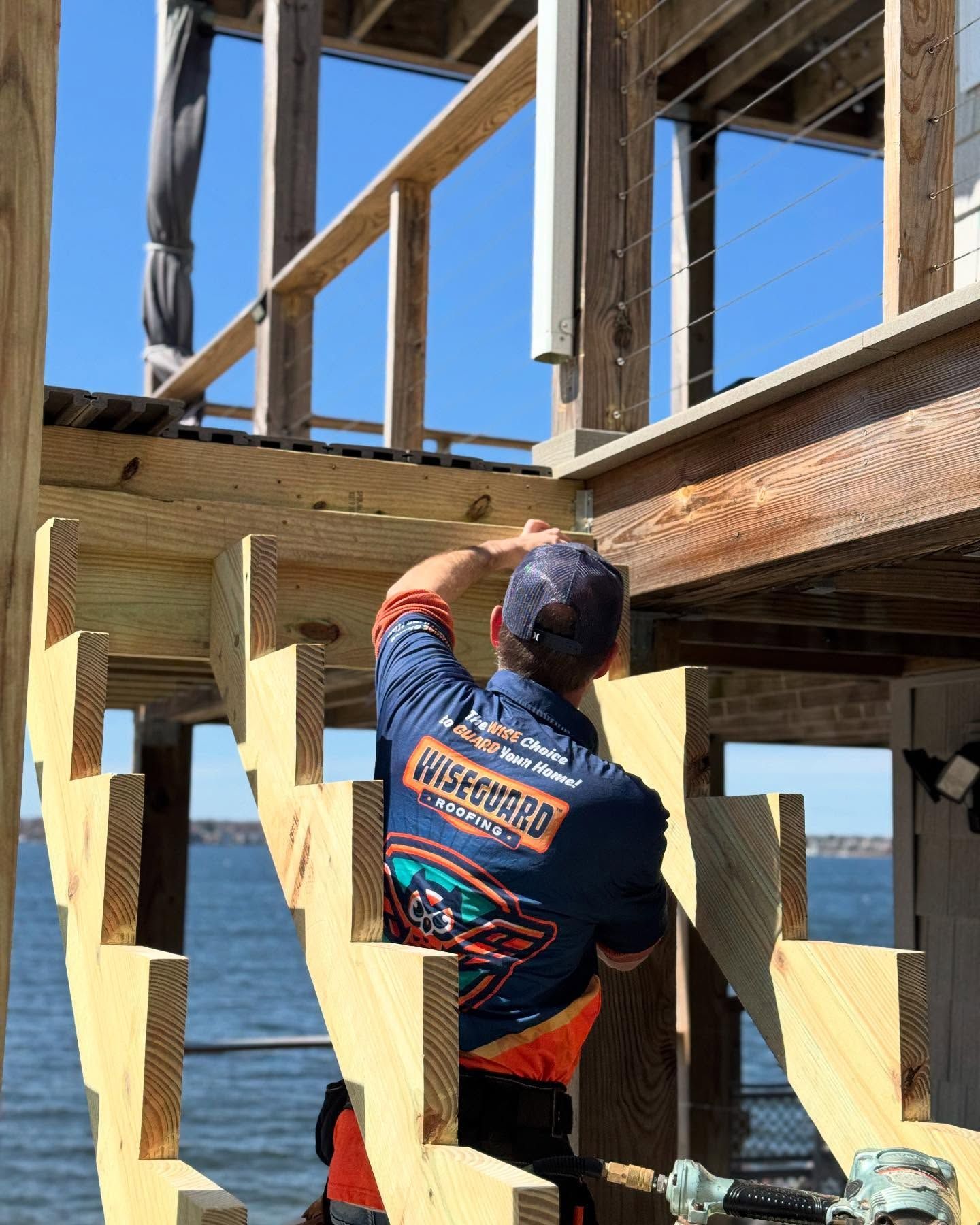 A worker in a dark blue uniform installs wooden stair stringers on a deck overlooking the water.