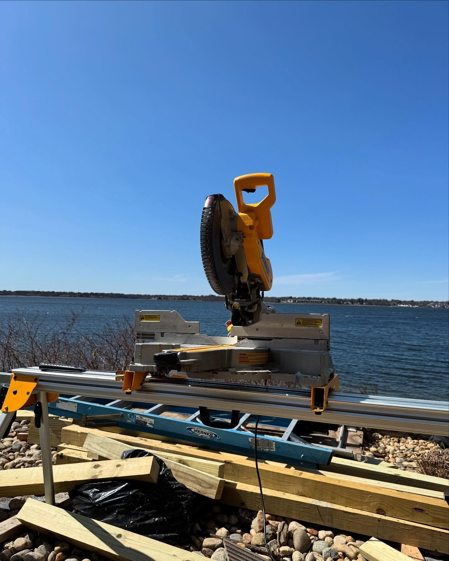 A yellow DeWalt miter saw mounted on a metal stand outdoors by a body of water under a clear blue sky.