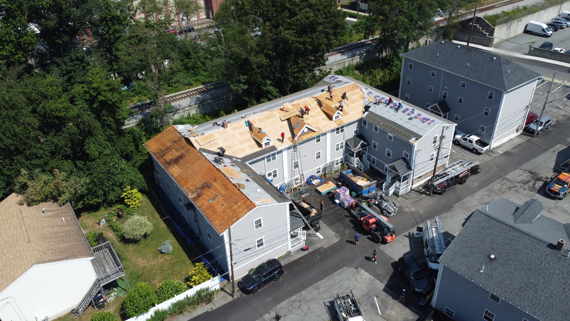 Aerial view of a partially demolished white building with exposed roof and debris, surrounded by houses and parked vehicles.