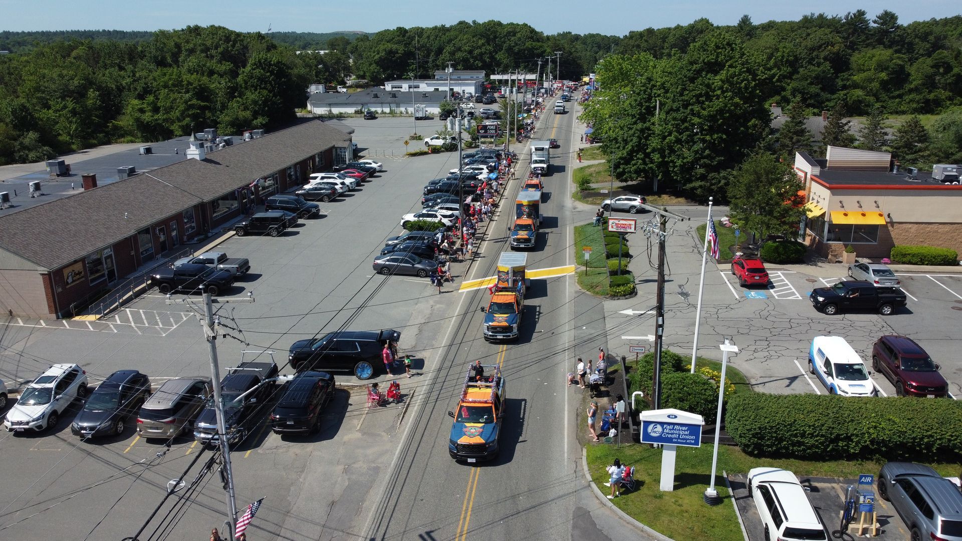 Aerial view of a busy parking lot with many cars, storefronts, and trees on a sunny day