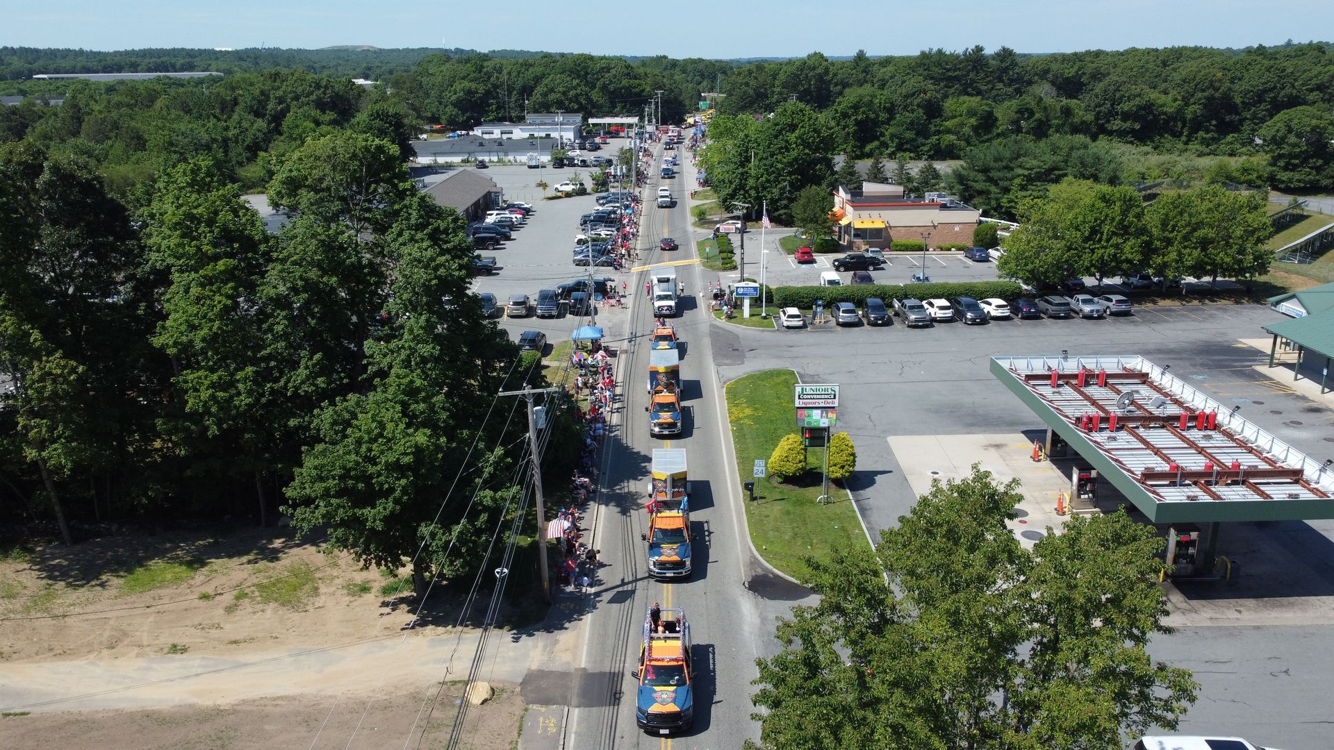 A long line of cars waits at an outdoor drive-thru beside a parking lot and trees.