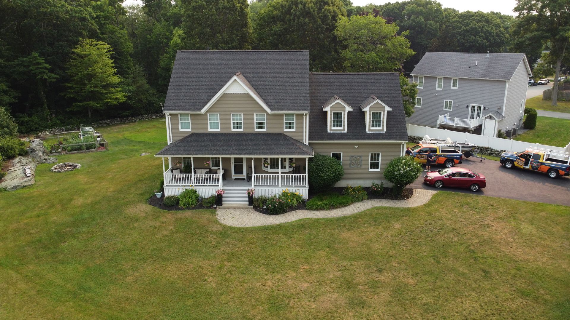 Aerial view of a gray two-story house with a front porch, large lawn, driveway, and parked cars.