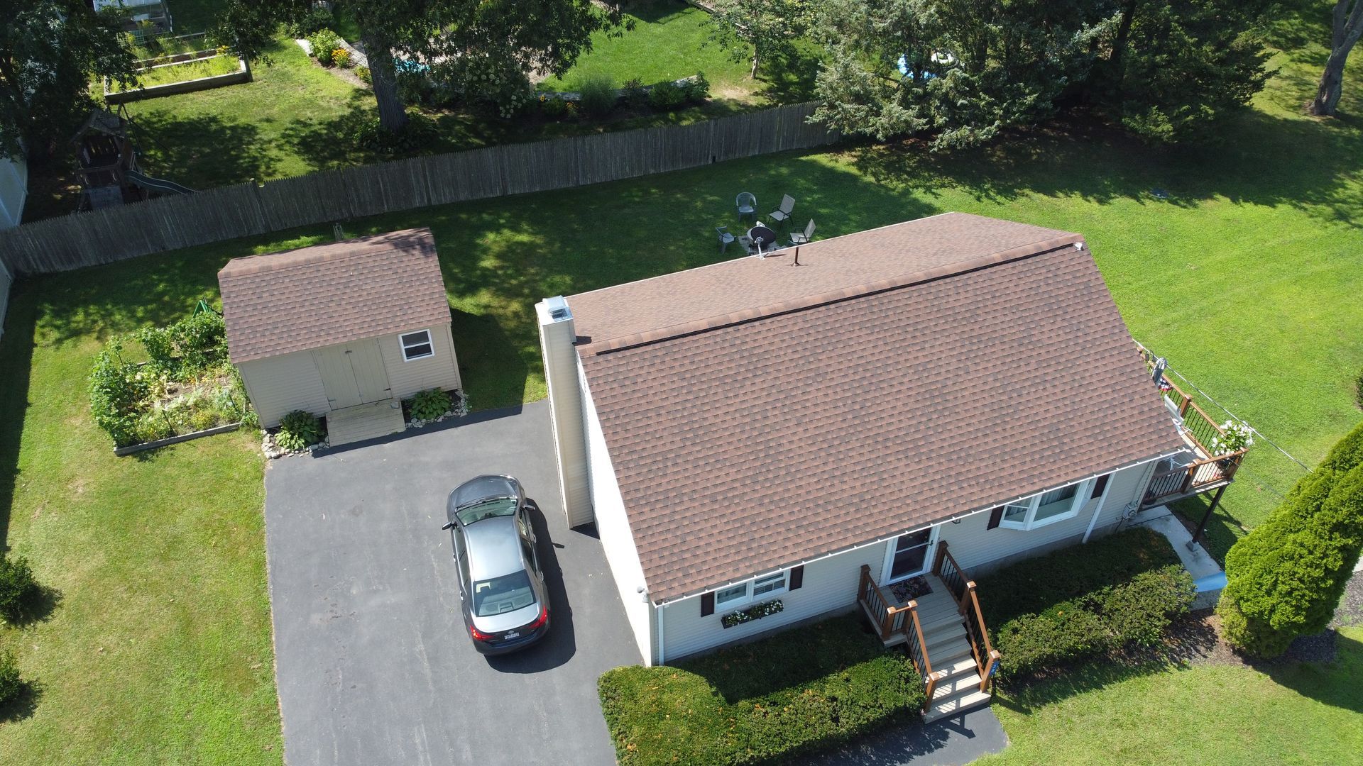 Aerial view of a white house with a brown roof, detached garage, driveway, and parked car surrounded by green lawn.