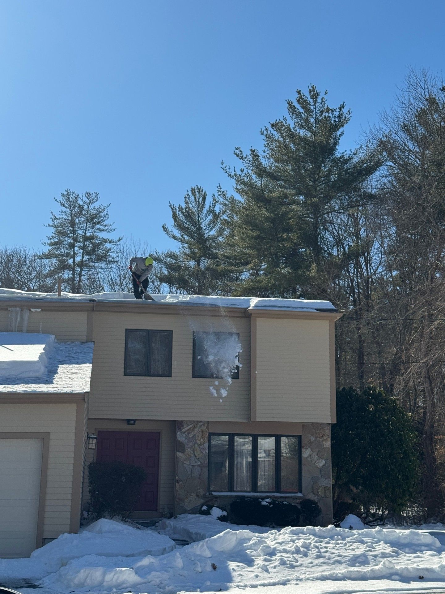 A person stands on the snow-covered roof of a two-story beige house, shoveling snow off the edge.