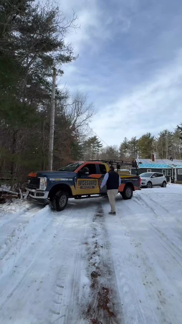 A person stands next to a service truck parked on a snowy road near trees and a house on a cloudy day.