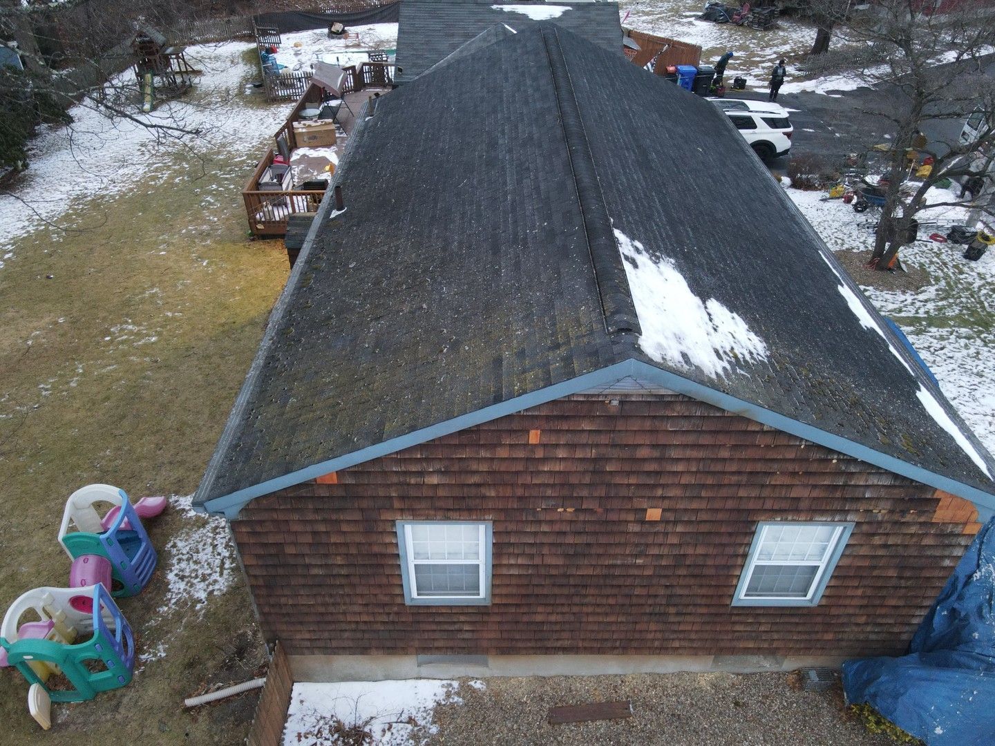 An aerial view of a brown shingled house with a dark roof partially covered in melting snow, next to a backyard.