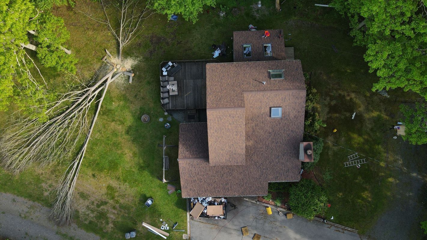An aerial view of a house with a brown shingled roof surrounded by trees and a fallen tree in the yard.