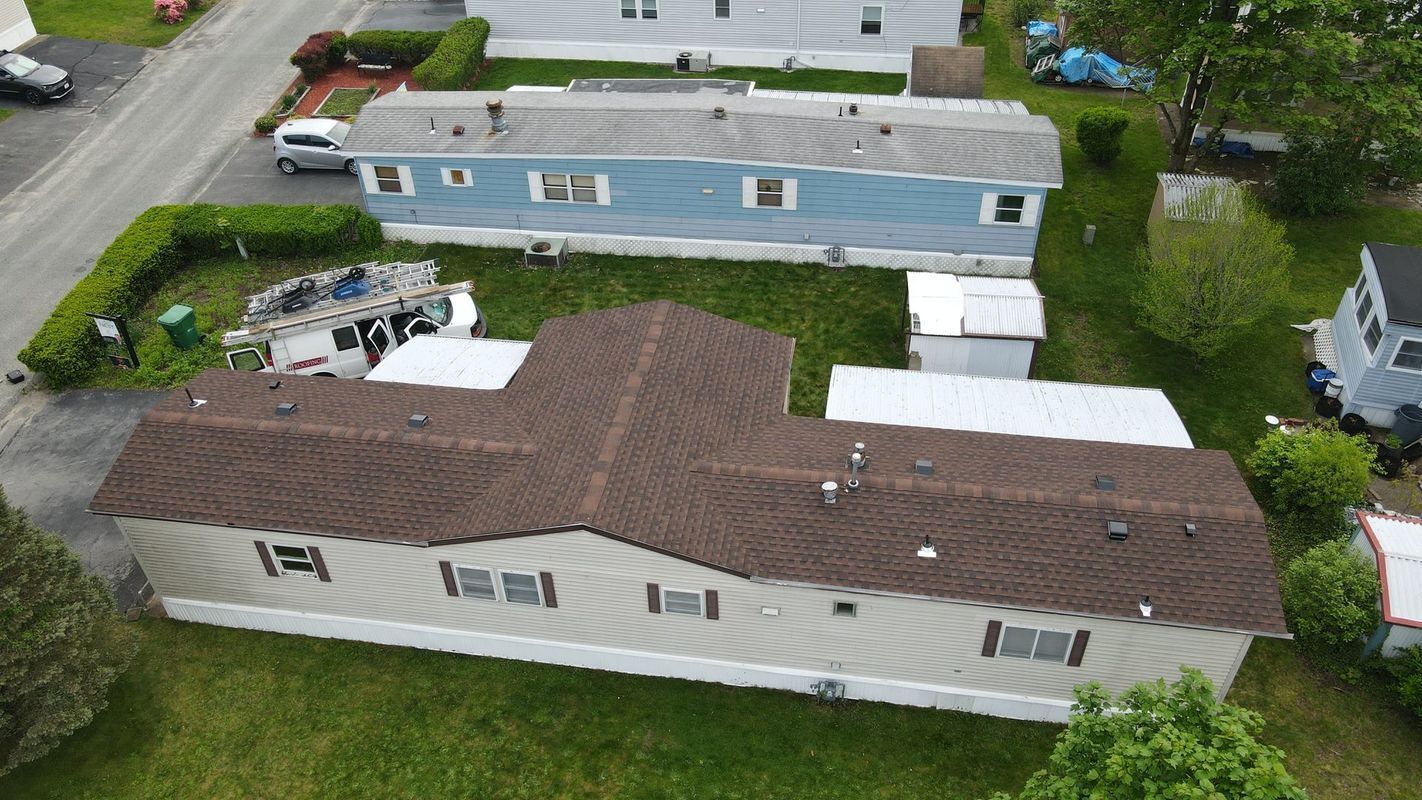 An elevated view of a tan mobile home with a dark brown shingled roof, next to a blue mobile home in a grassy lot.
