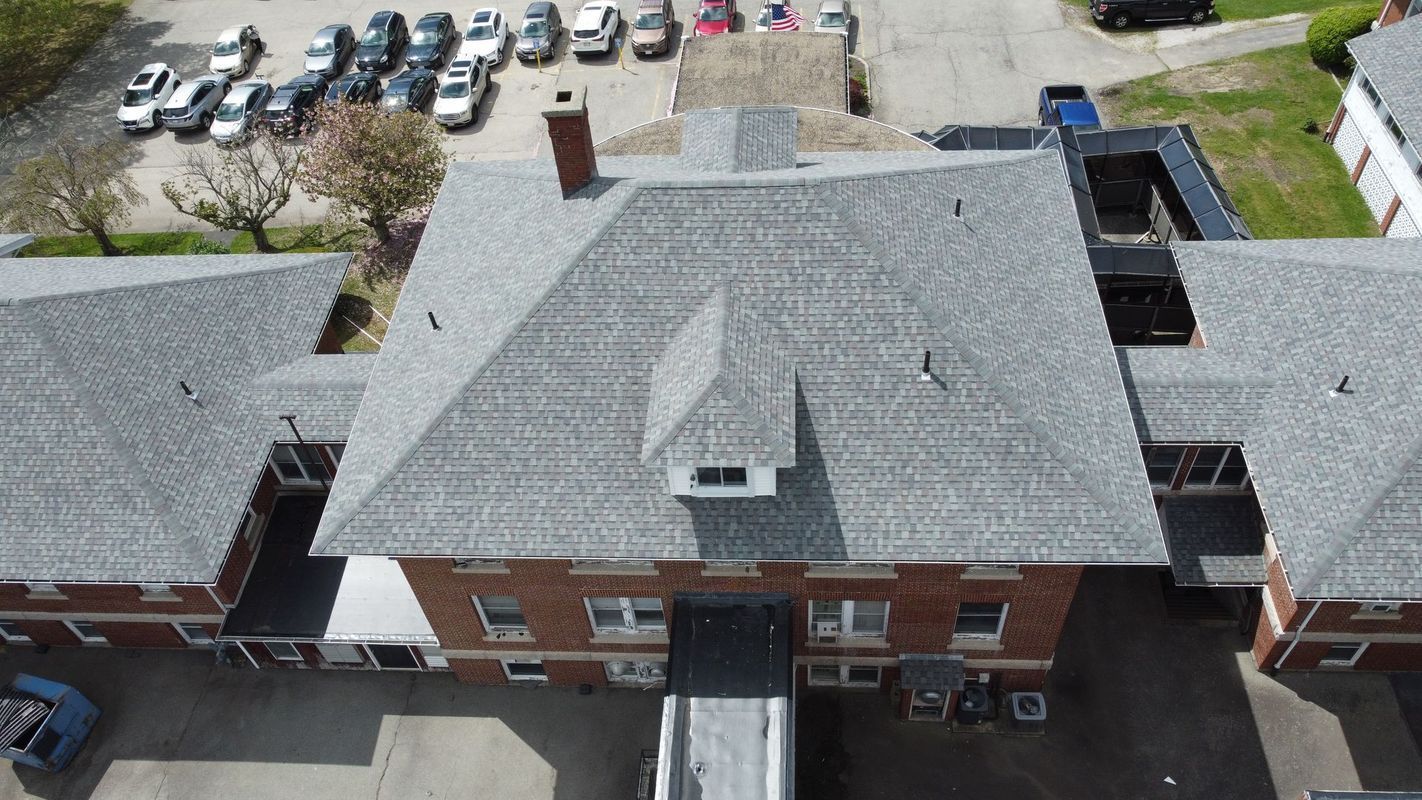 Aerial view of a brick building with a gray shingled roof, a chimney, and a small dormer, adjacent to a paved parking lot.