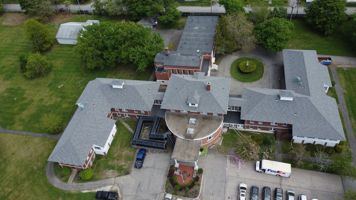 Aerial view of a sprawling brick building with a gray shingled roof, surrounding grounds, and a parking lot.