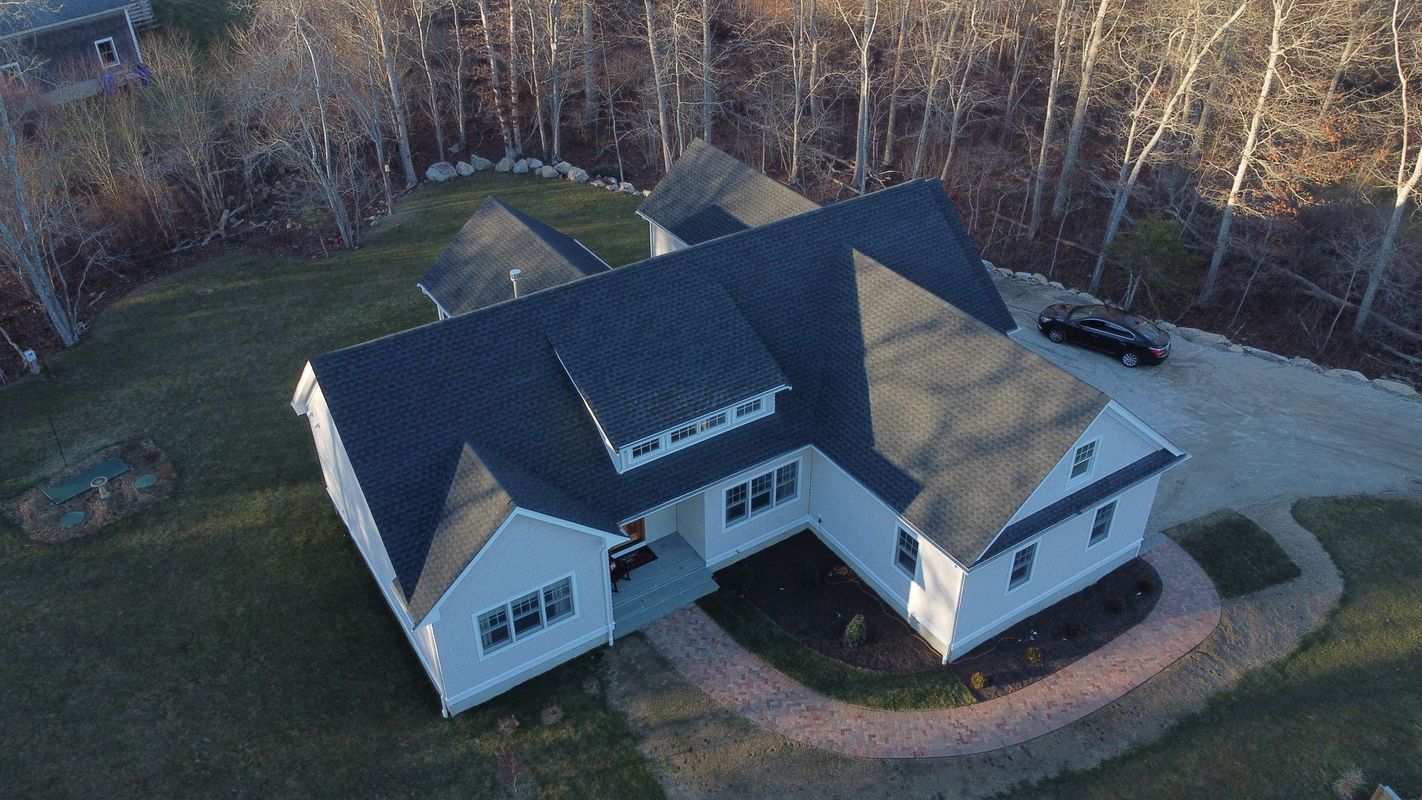 An aerial view of a white, modern house with a dark metal roof, set on a grassy lawn with a curved brick pathway.