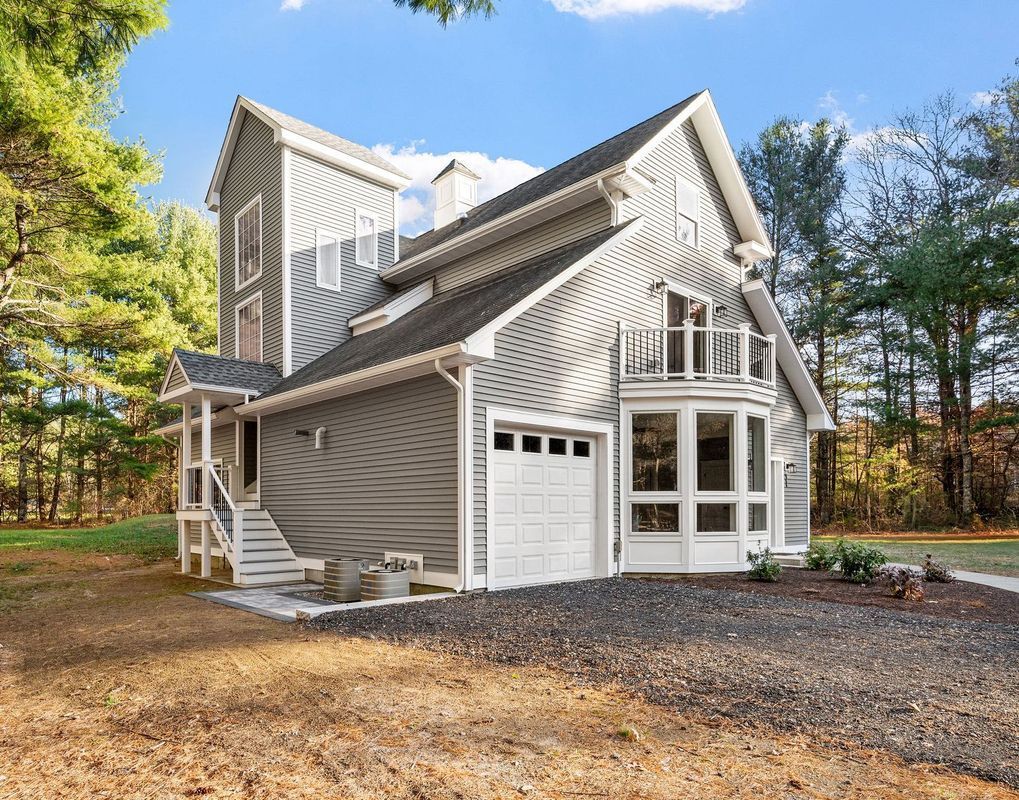 A two-story grey, contemporary house with a steep roof, garage, and small balcony, situated in a wooded area.