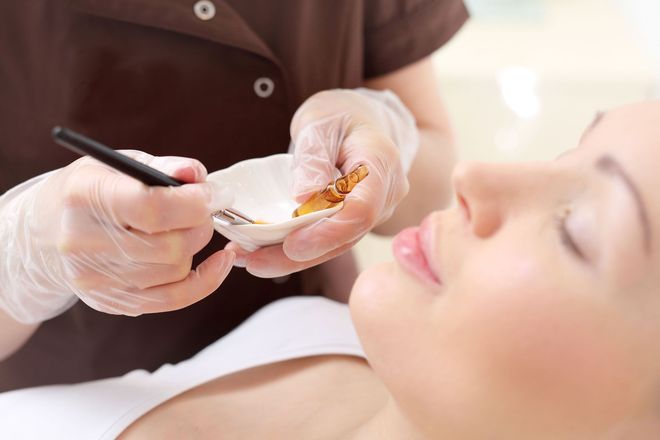 A person receiving a facial treatment. A gloved hand holds a brush and bowl near the woman's face.