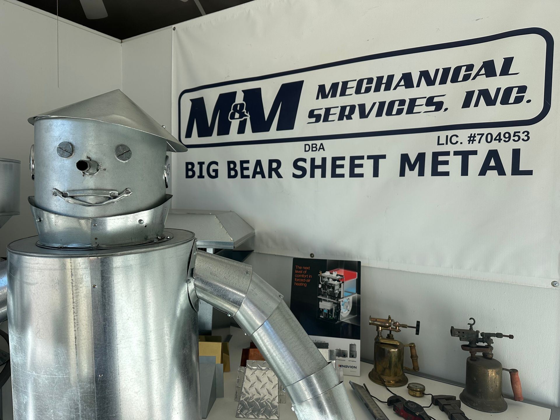 A metal robot figure stands in front of an M&M Mechanical Services, Inc. banner in a workshop setting.