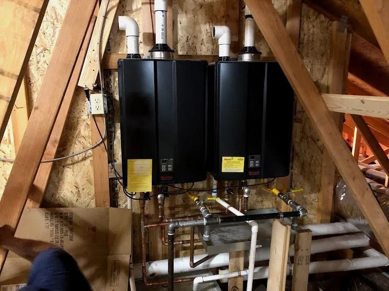 Two black tankless water heaters mounted on an attic wall with white PVC venting and copper plumbing connections.