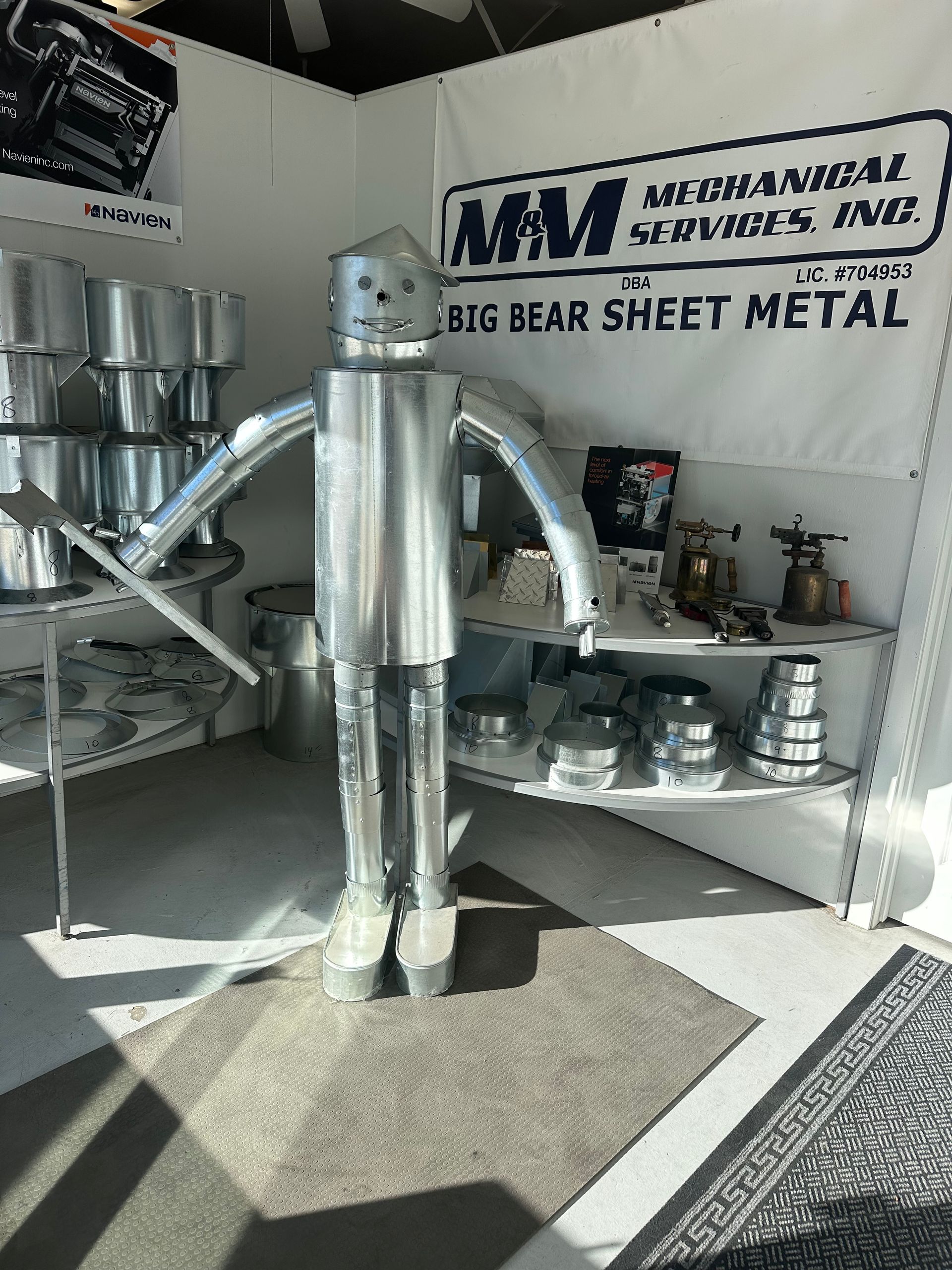 A metal robot sculpture made of sheet metal parts stands in a workshop featuring a banner for a mechanical company.