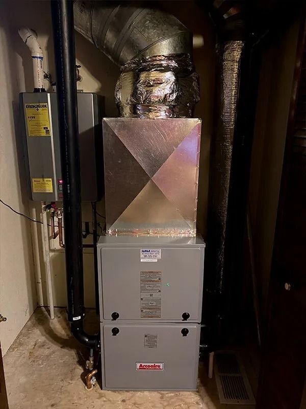 A gray gas furnace with a metal duct above and a tankless water heater on the left in a utility closet.