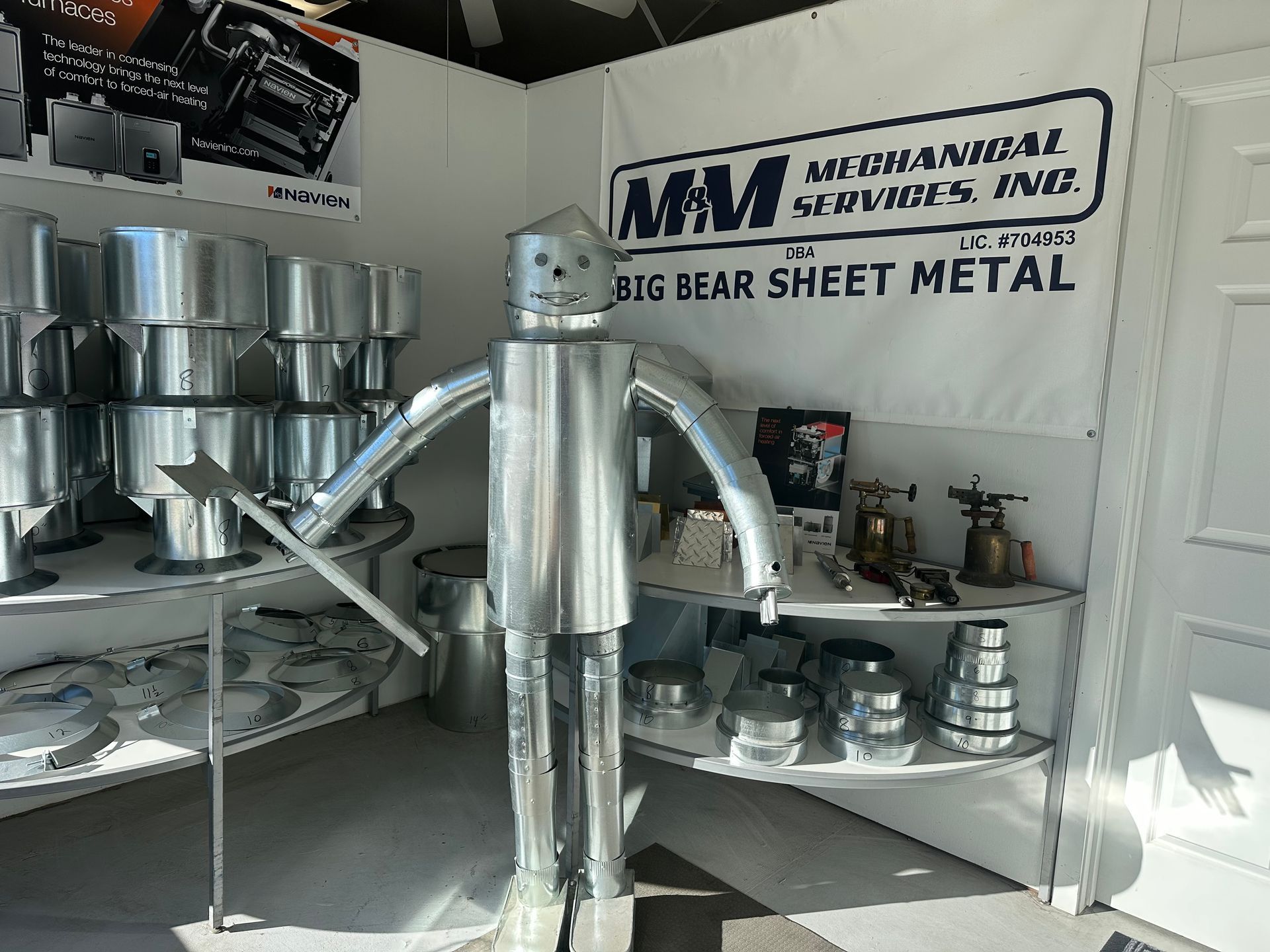 A tin robot sculpture stands in the center of a sheet metal workshop with shelving units and a company sign in the back.
