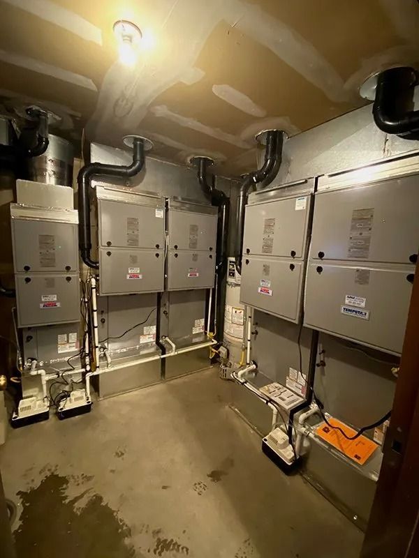 A utility room containing four large grey HVAC furnace units with attached black vent pipes and white drainage lines.