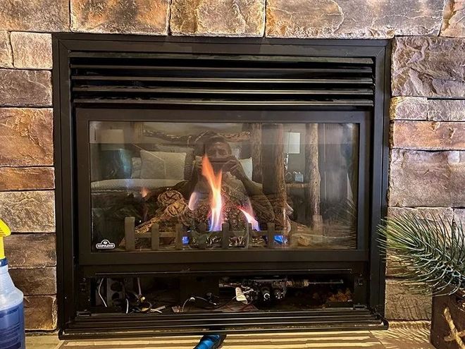 A glowing gas fireplace set into a rustic stone wall, with the bottom compartment open to reveal internal components.