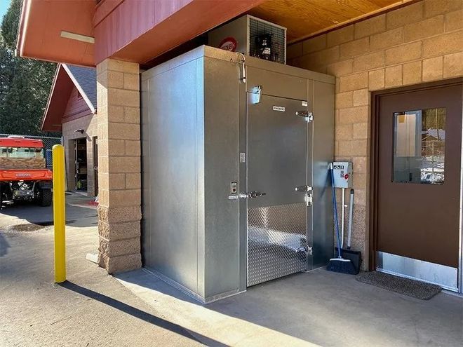 A metal walk-in cooler stands under a building overhang next to a brown door, with a utility vehicle parked nearby.