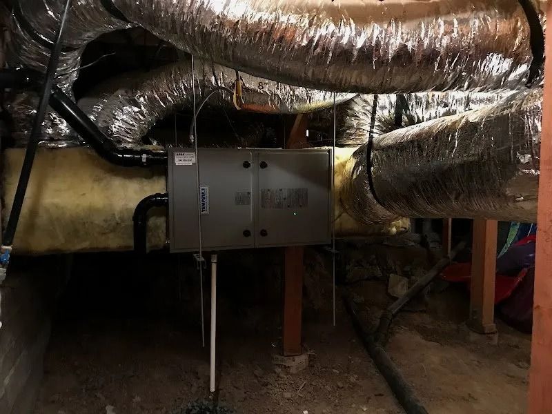 A gray HVAC air handler unit suspended in a crawl space, connected to insulated silver ductwork and drainage pipes.