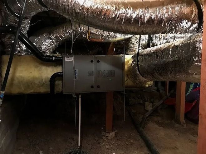 A grey HVAC air handler unit suspended in a crawlspace, connected to flexible silver air ducts and insulation.