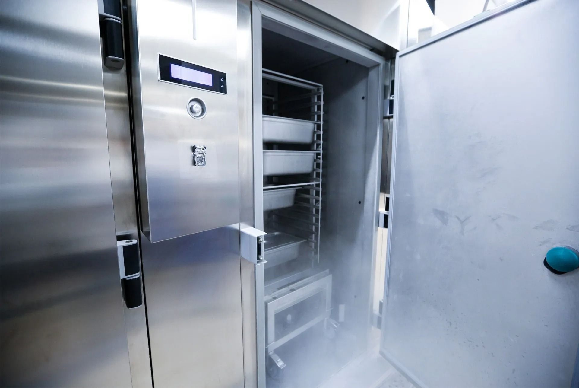 A stainless steel commercial blast chiller or freezer with its door open, emitting cold mist over internal food racks.