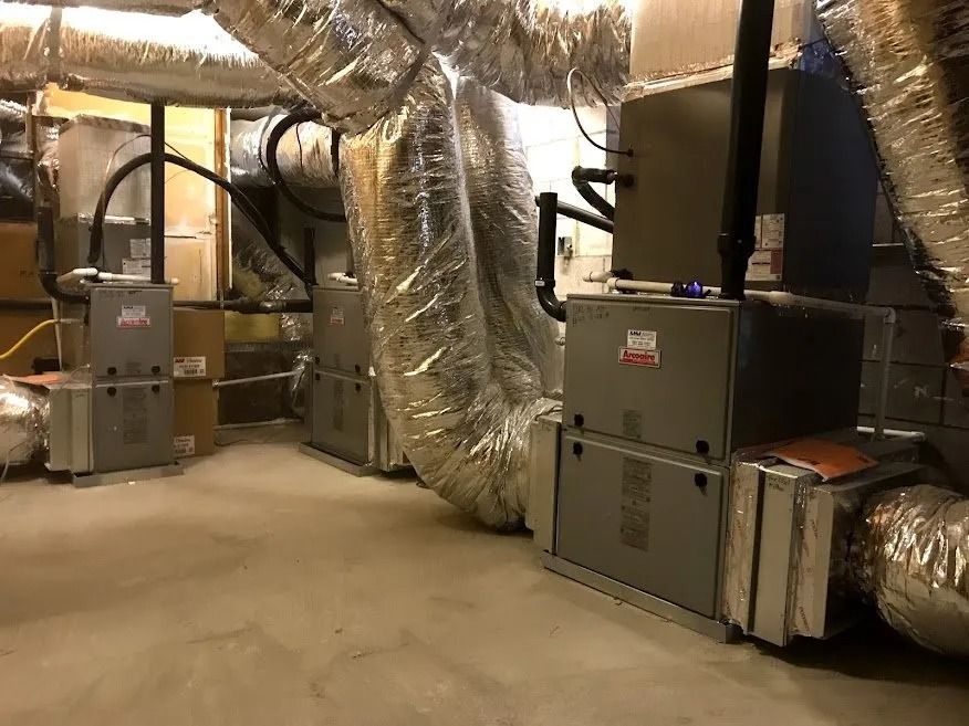 A basement with three grey HVAC furnace units connected to large, silver metallic insulated ductwork.