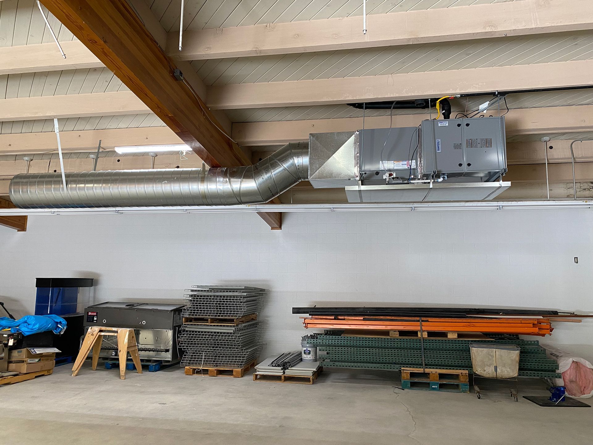 An industrial HVAC unit suspended from wooden ceiling beams above stacked materials in a storage area.