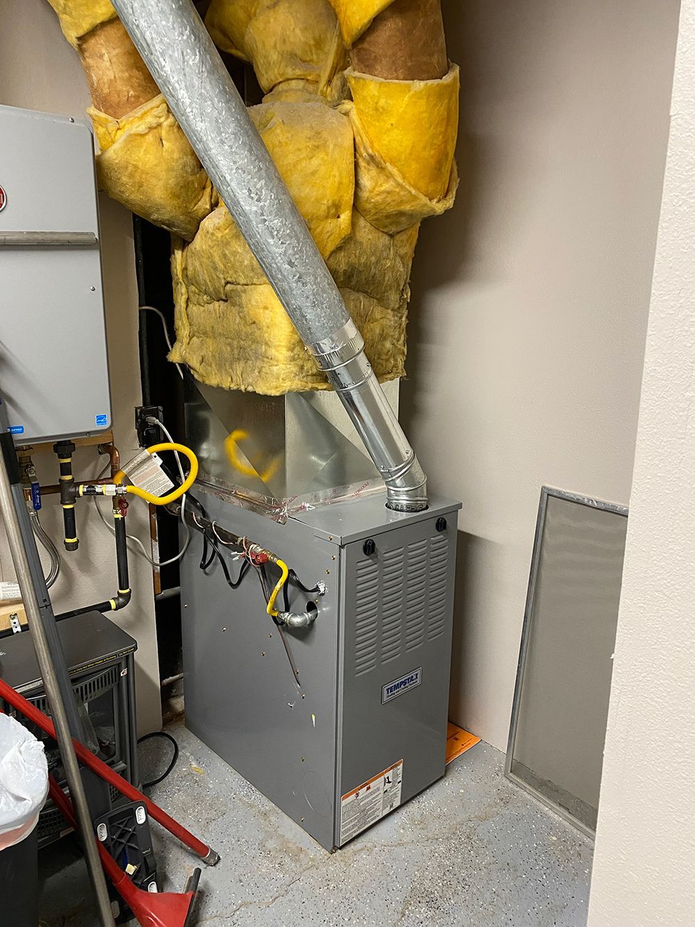 A gray HVAC furnace with a metallic exhaust pipe stands in a corner with yellow insulation visible above it.