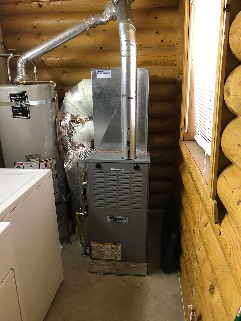A gray furnace and a white water heater are installed in a corner of a room with log cabin walls.