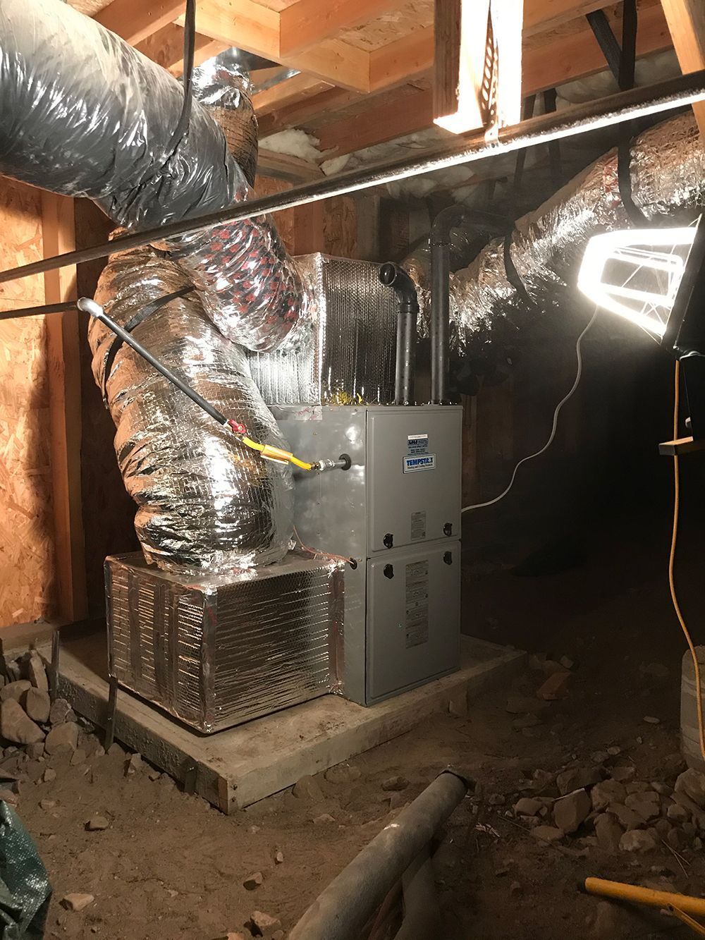 A gray HVAC unit sits on a concrete pad in a crawlspace, connected to silver insulated ducts under wooden beams.