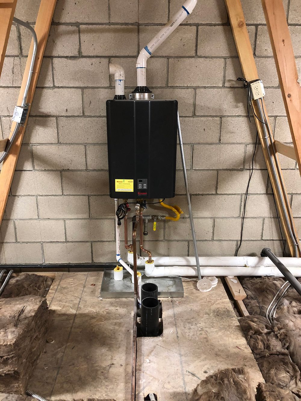 A black tankless water heater mounted on a concrete block wall in an attic with associated piping and exhaust venting.