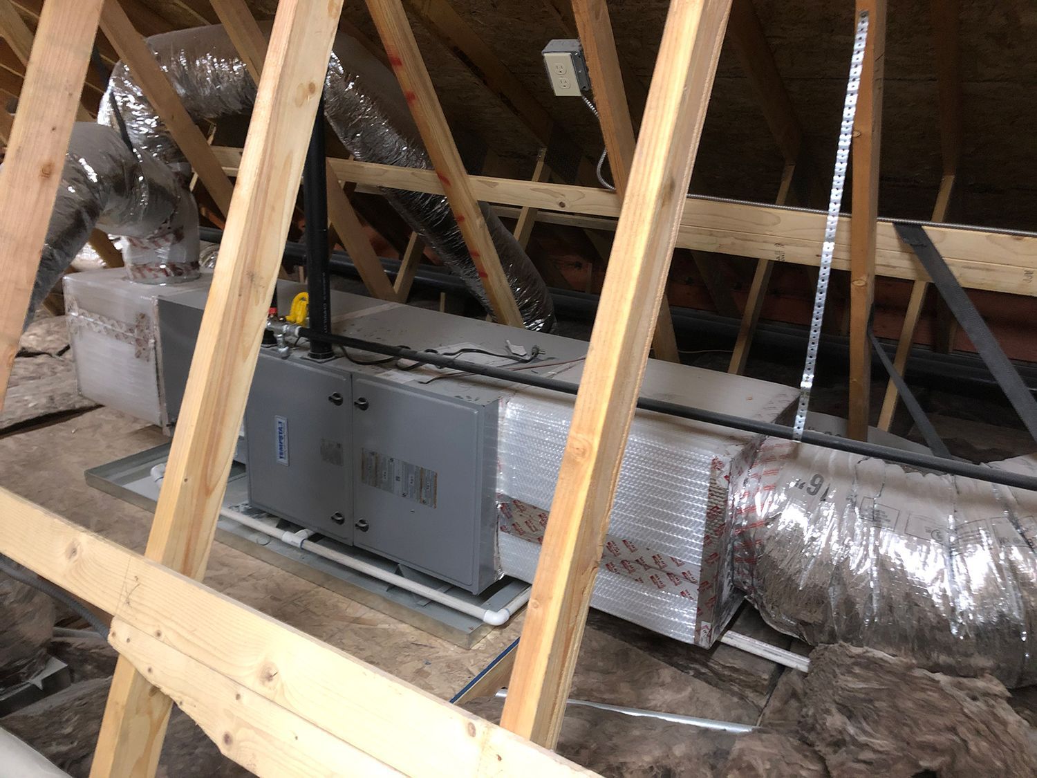 An HVAC air handler unit with insulated ductwork installed in an attic space among wooden rafters.