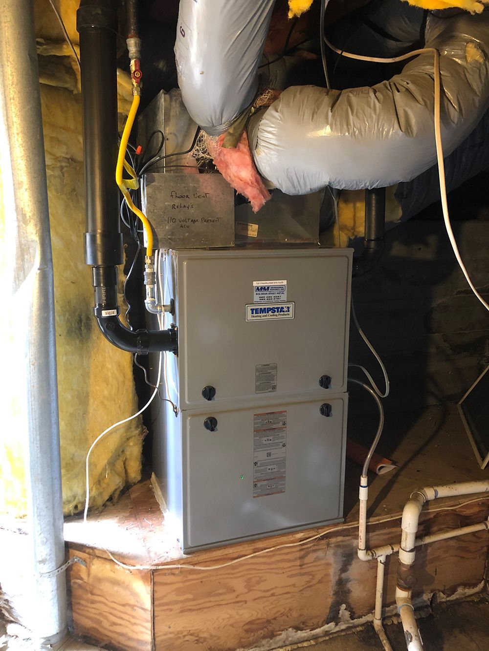 A gray HVAC furnace unit is installed in a basement, connected to silver ductwork and PVC venting pipes.