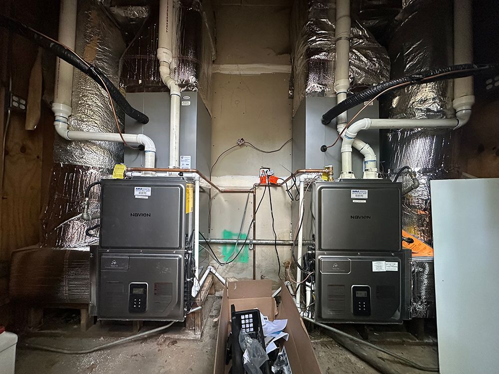 Two modern dark gray HVAC units stand side-by-side in a utility room with exposed ductwork and white PVC pipes.