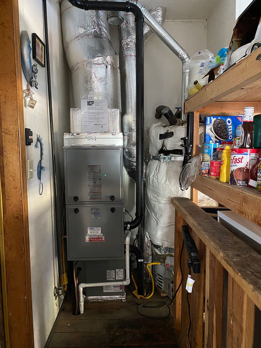 A utility closet with a grey furnace and insulated piping, situated beside a wooden shelf holding various food items.