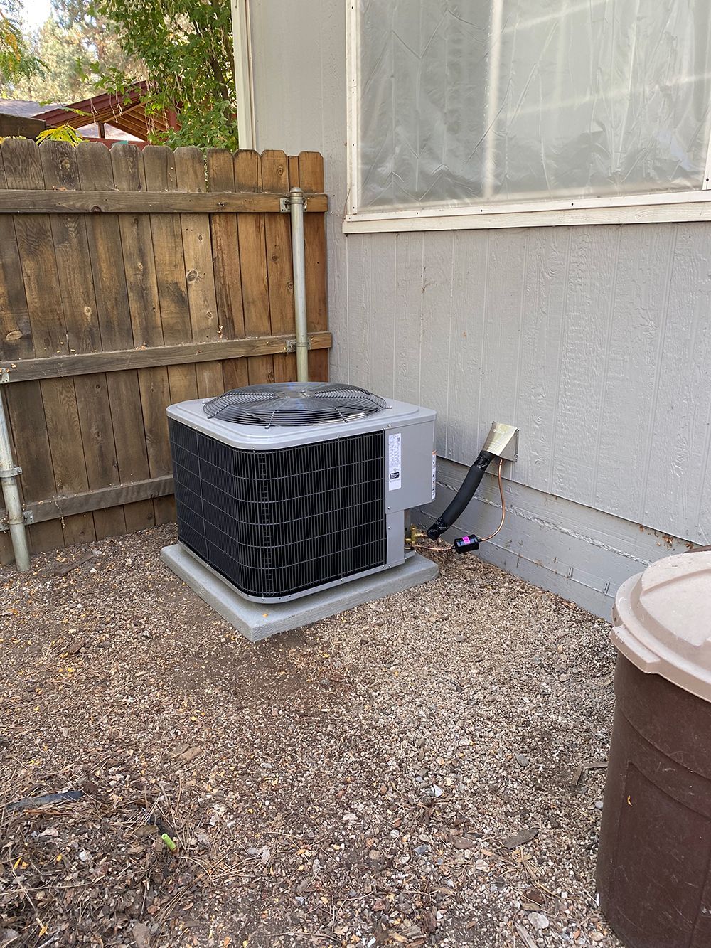 An outdoor air conditioning unit sits on a gravel patch beside a house wall and a wooden fence.