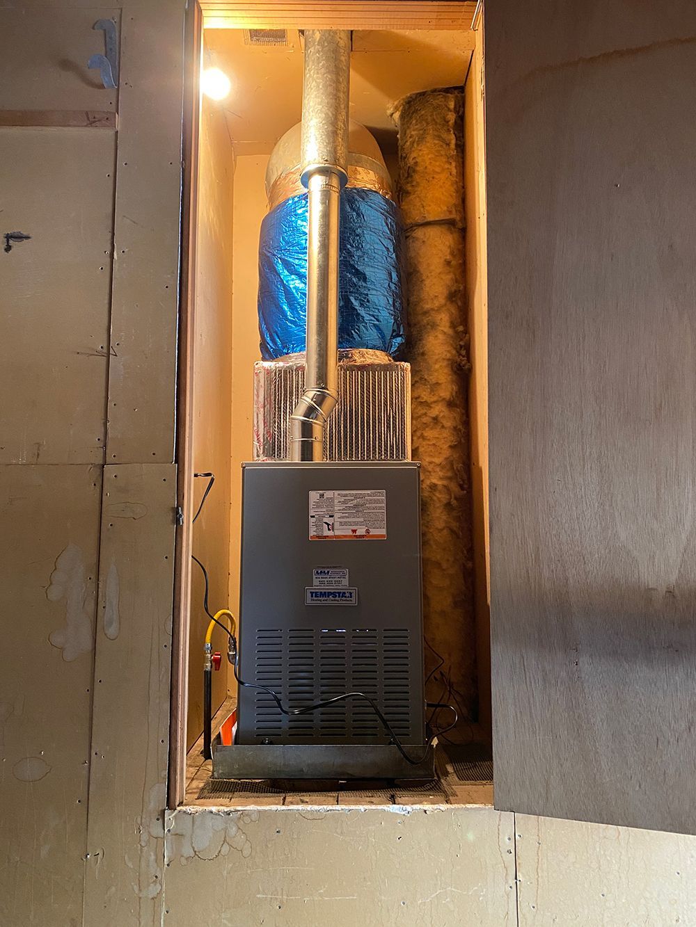 A gray gas furnace installed in a narrow closet with blue-wrapped ductwork and a vertical chimney pipe extending upward.