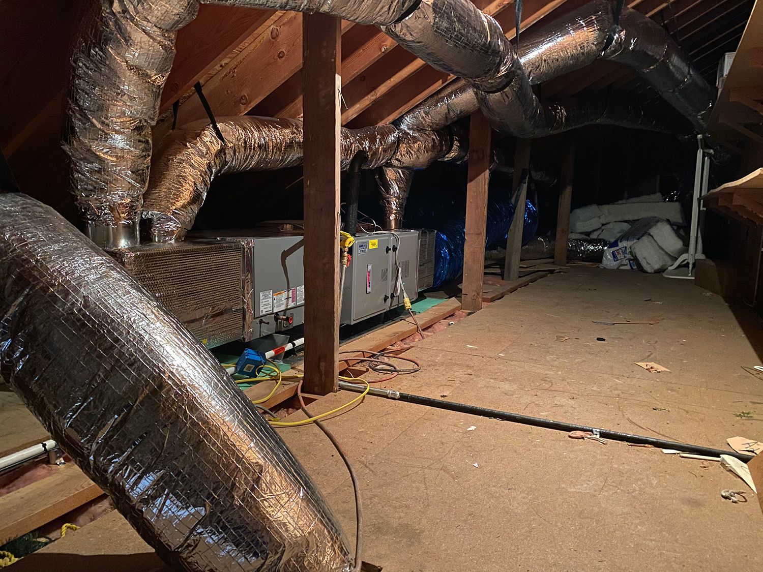 A central air conditioning unit with metallic flexible ductwork sits on the floor of an unfinished wooden attic.