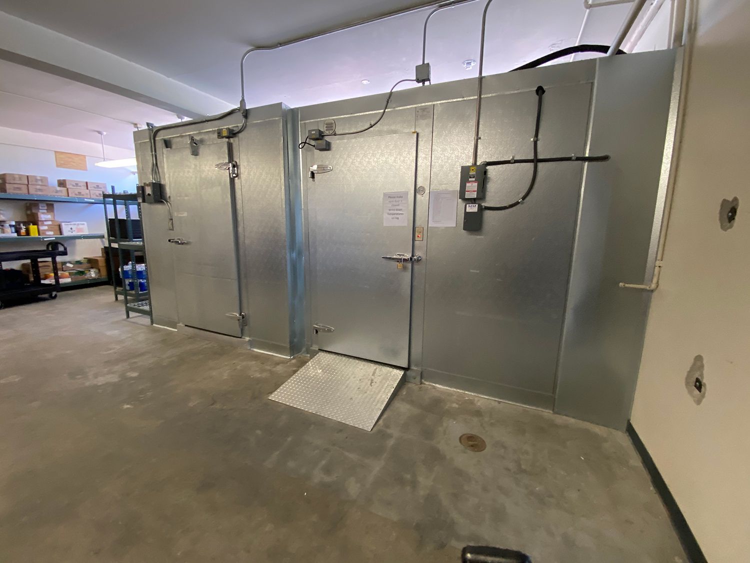 Two industrial walk-in coolers with metal doors in a room with a concrete floor and shelving in the background.