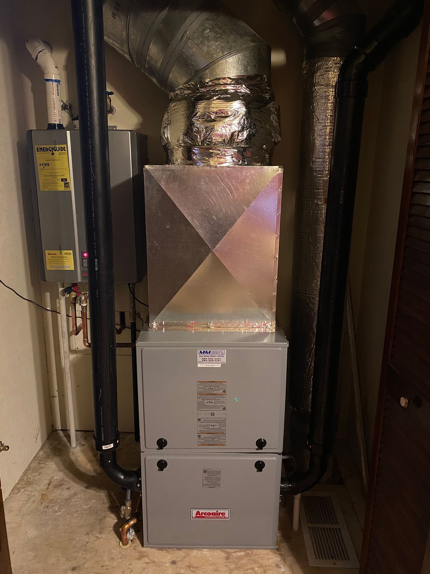 A grey HVAC furnace unit with a metal duct above and black pipes on either side, situated in a utility closet.