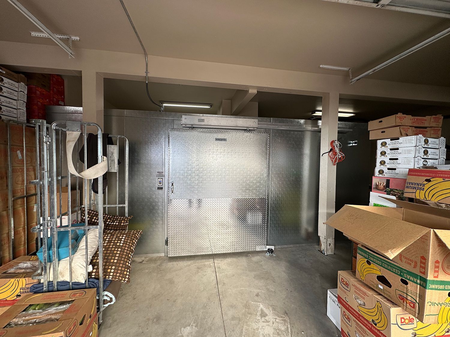 A walk-in cooler with metal doors sits in a storage area next to stacks of cardboard boxes and a rolling metal cart.