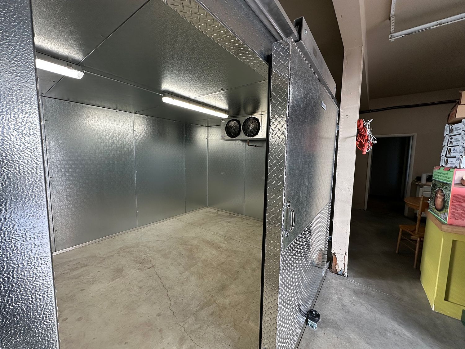 An open, walk-in industrial freezer with metallic, textured walls and ceiling, illuminated by two internal light fixtures.