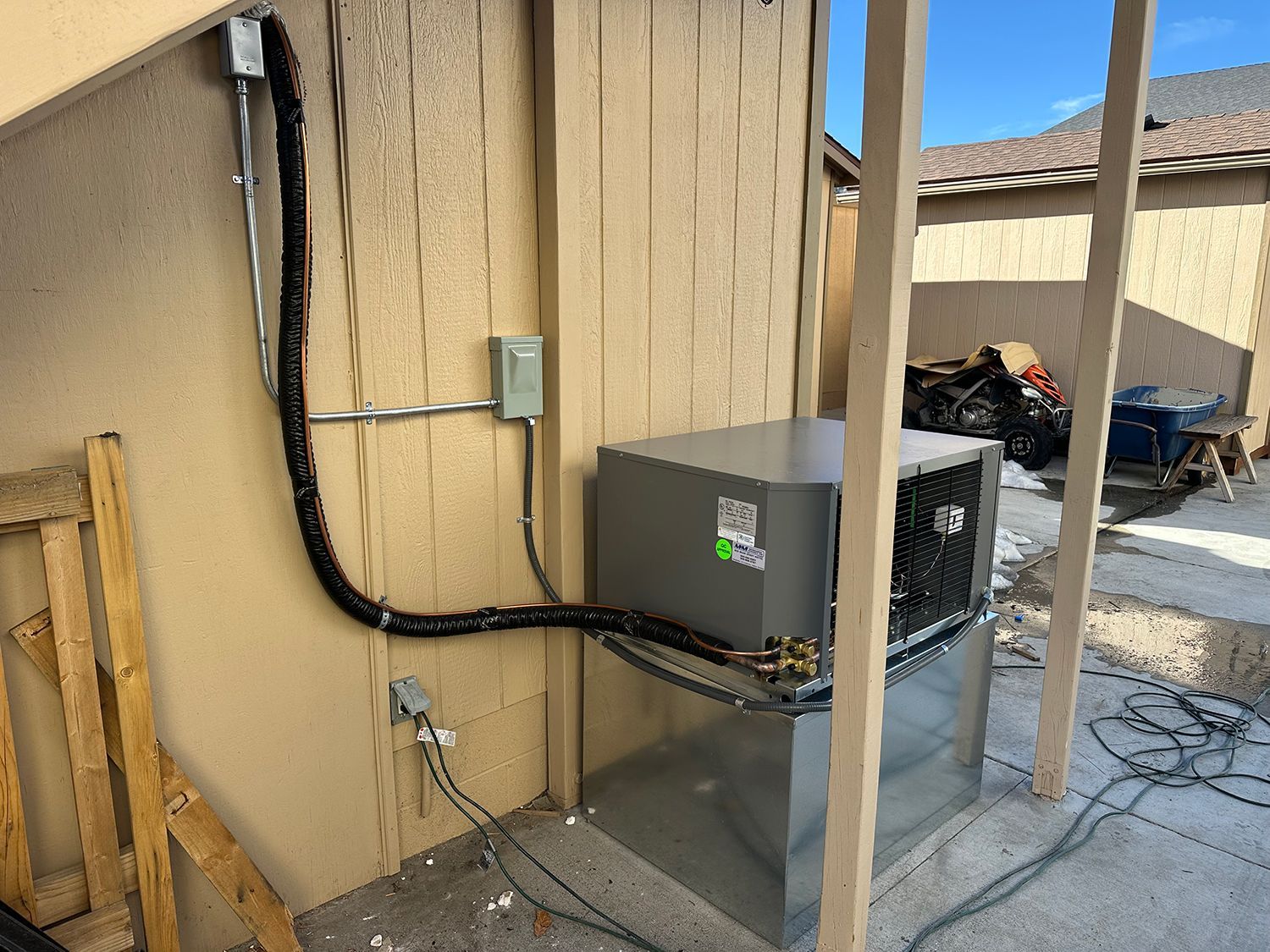 A gray air conditioning unit sits on a metal stand against a beige house wall with exposed electrical and pipe connections.