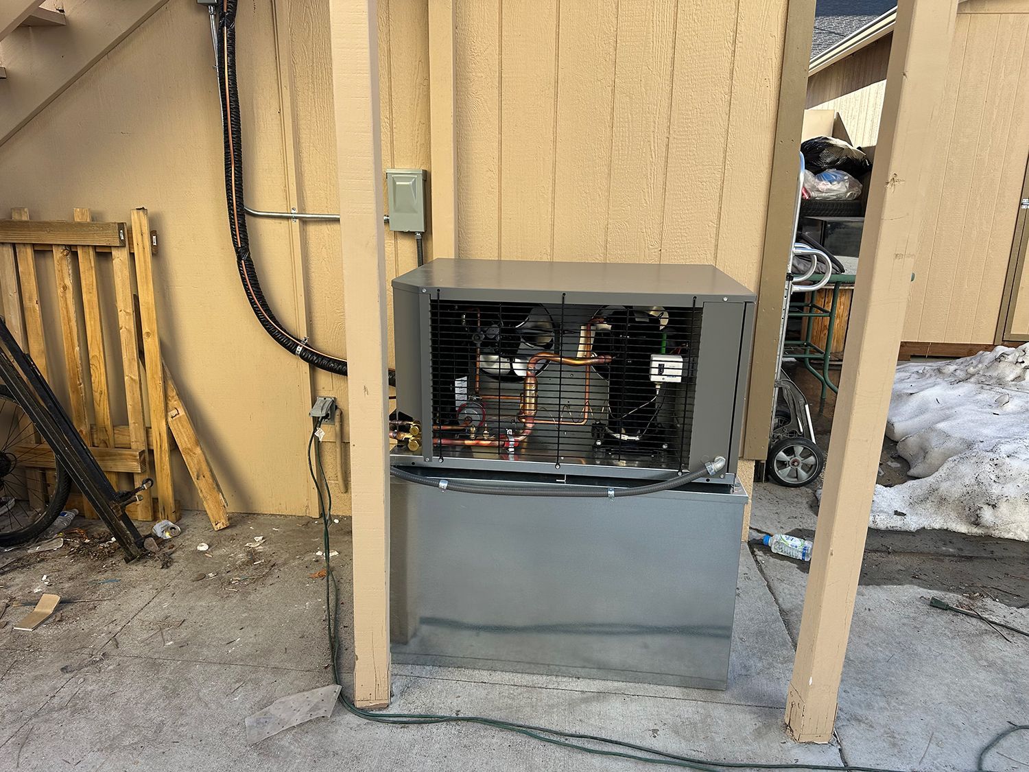 An outdoor air conditioning unit with an exposed internal panel, mounted against a tan wall under a wooden structure.