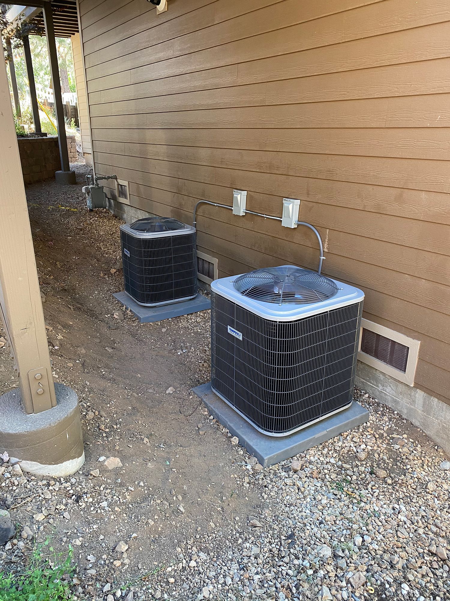 Two HVAC outdoor condenser units sit on gray pads against the side of a brown-sided building with rock ground cover.