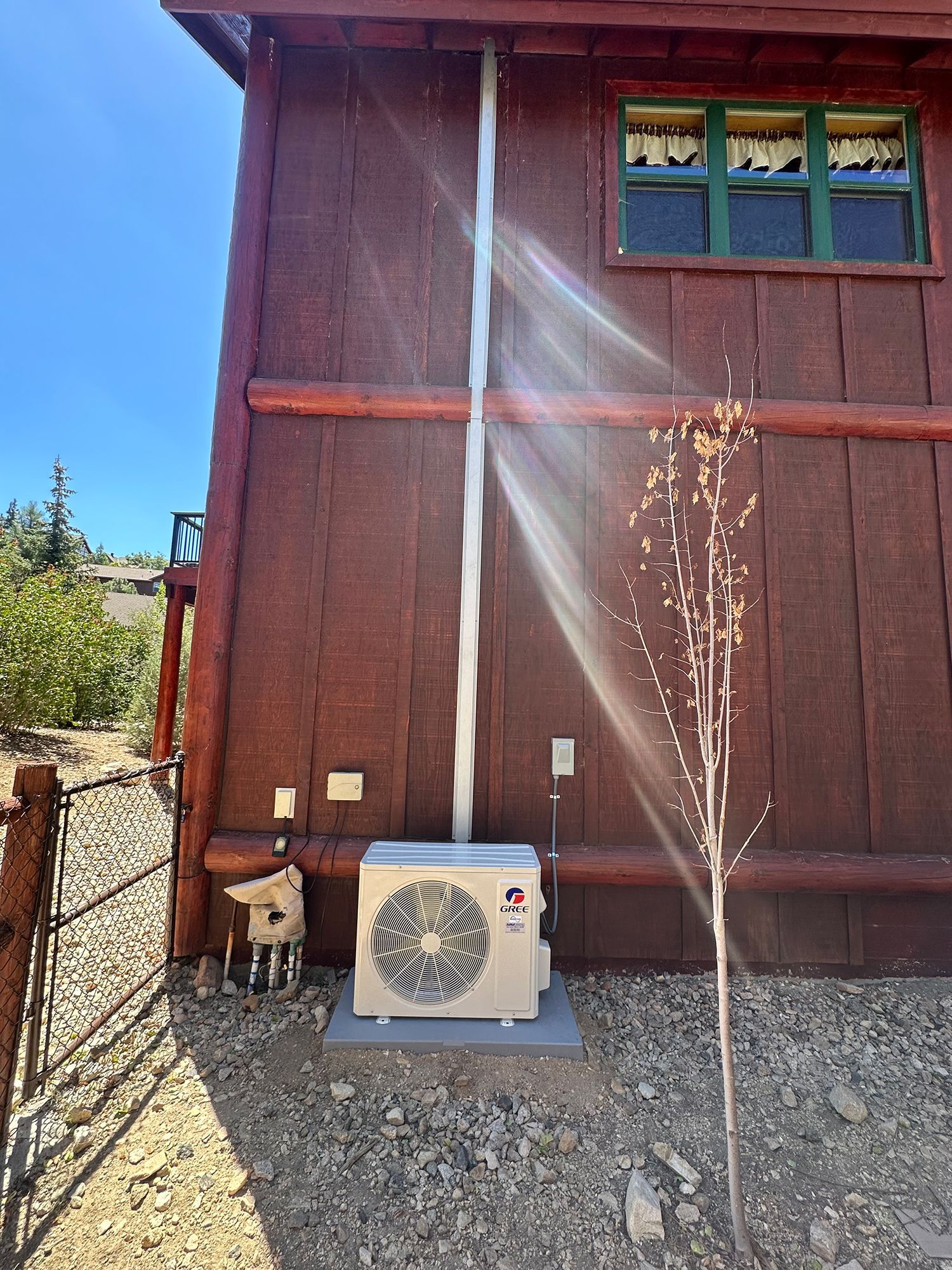 A tan outdoor heat pump unit sits against a rustic dark wood wall below a vertical white conduit pipe under a window.